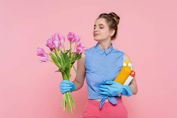 Piaciuta giovane donna in guanti di gomma che tiene bottiglie con detergente e tulipani odorosi isolati su rosa — Foto stock