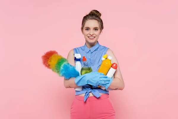 Mujer joven y complacida con guantes de goma sosteniendo botellas con detergente y cepillo de polvo aislado en rosa - foto de stock