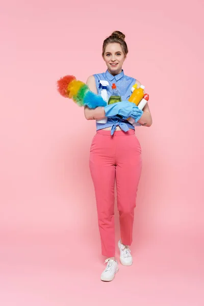 Pleine longueur de jeune femme heureuse dans des gants en caoutchouc tenant des bouteilles avec détergent et brosse à poussière sur rose — Photo de stock