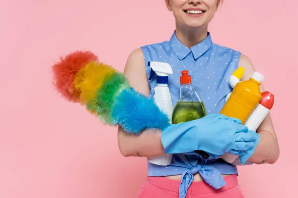 Cropped view of pleased young woman in rubber gloves holding bottles with detergent and dust brush isolated on pink — Stock Photo
