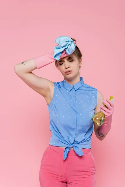 Mujer joven y cansada con guantes de goma sosteniendo trapo azul y botella de spray aislado en rosa - foto de stock