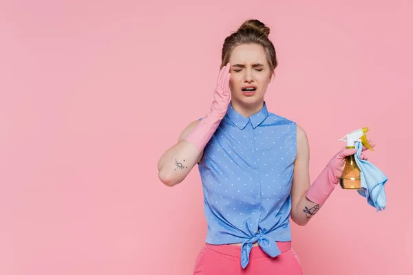 Exhausted young woman in rubber gloves holding blue rag and spray bottle isolated on pink — Stock Photo