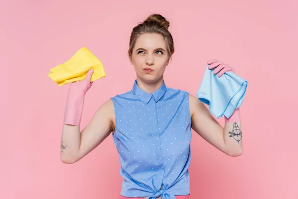 Nachdenkliche junge Frau in Gummihandschuhen mit blauen und gelben Lumpen auf rosa — Stockfoto