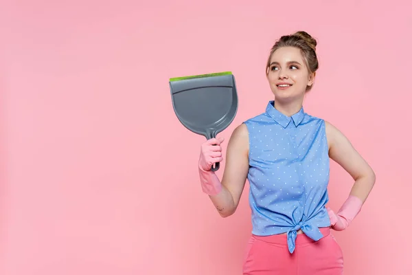 Glückliche junge Frau in Gummihandschuhen, die Kehrschaufel hält und mit der Hand an der Hüfte isoliert auf rosa steht — Stockfoto