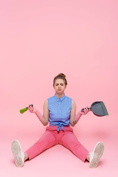 Full length of sad young woman in rubber gloves holding broom and dustpan while sitting on pink — Stock Photo