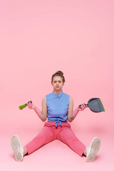 Longitud completa de la triste mujer joven en guantes de goma que sostiene la escoba y el recogedor mientras está sentado en rosa - foto de stock