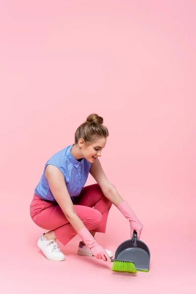 Pleine longueur de jeune femme heureuse dans des gants en caoutchouc tenant balai et poêle à poussière tout en faisant le nettoyage de la maison sur rose — Photo de stock