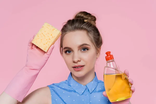 Mujer joven sosteniendo esponja y botella con detergente aislado en rosa - foto de stock