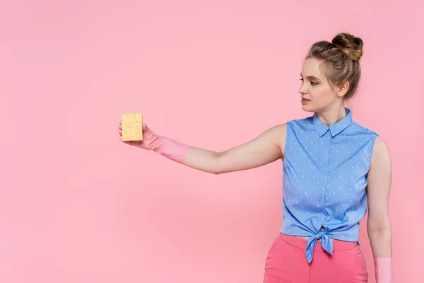 Junge Frau in Gummihandschuhen blickt auf Schwamm isoliert auf rosa — Stockfoto