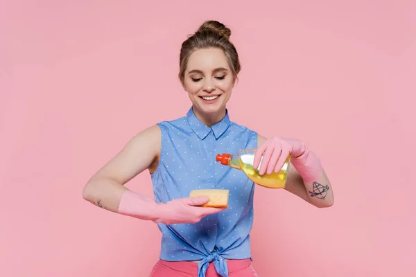 Mujer joven tatuada sosteniendo esponja y botella con detergente mientras sonríe aislada en rosa - foto de stock