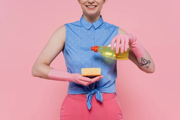 Visión parcial de mujer feliz y tatuada vertiendo detergente sobre esponja aislada en rosa - foto de stock