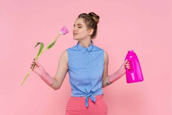 Young woman holding bottle with detergent and smelling tulip isolated on pink — Stock Photo