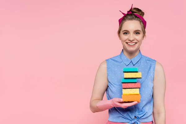 Feliz joven con guantes de goma sosteniendo esponjas apiladas aisladas en rosa - foto de stock