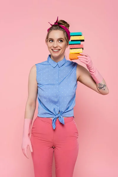 Joyful young woman in rubber gloves holding stacked sponges isolated on pink — Stock Photo