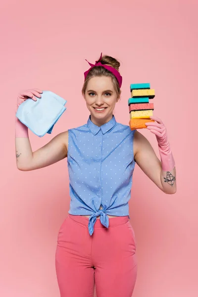 Feliz joven con guantes de goma sosteniendo esponjas apiladas y trapo aislado en rosa - foto de stock