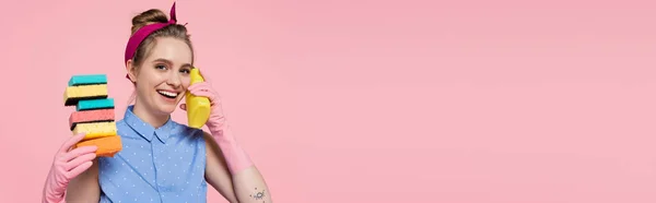 Jovem feliz em luvas de borracha segurando esponjas empilhadas e garrafa isolada em rosa, banner — Fotografia de Stock