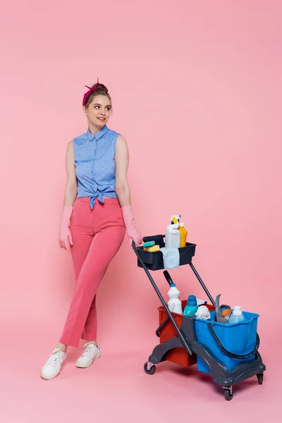 Volle Länge der lächelnden jungen Frau in Gummihandschuhen, die in der Nähe des Hauswirtschaftswagens mit Putzmitteln auf rosa steht — Stockfoto