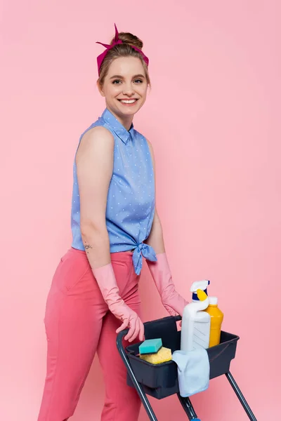 Felice giovane donna in guanti di gomma in piedi vicino al carrello delle pulizie con prodotti per la pulizia isolati su rosa — Foto stock