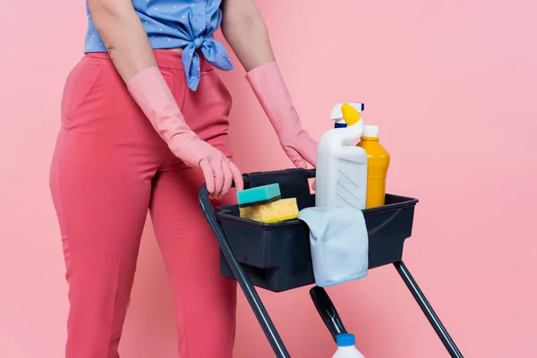 Vista ritagliata di donna tatuata in guanti di gomma in piedi vicino al carrello di pulizia con detergenti isolati su rosa — Foto stock