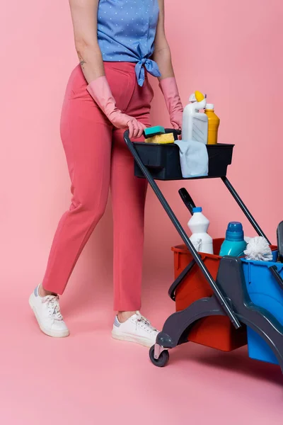 Vue recadrée de la femme tatouée dans des gants en caoutchouc debout près du chariot d'entretien ménager avec des fournitures de nettoyage sur rose — Photo de stock