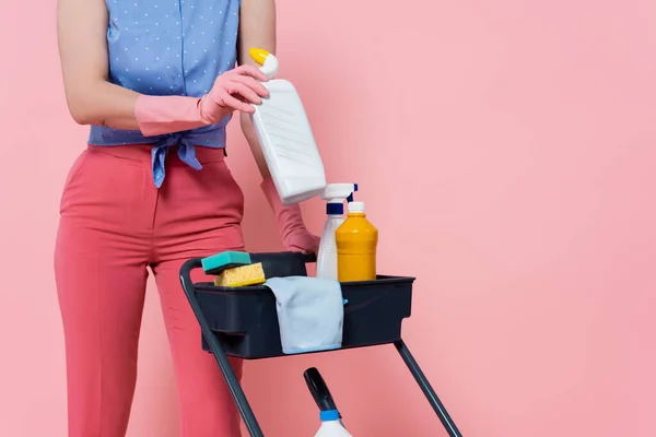 Cropped view of tattooed woman in rubber gloves standing near housekeeping cart and holding bottle isolated on pink — Stock Photo