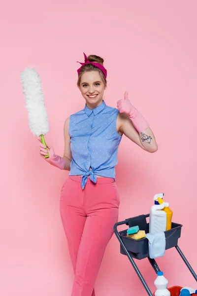 Cheerful young woman standing near housekeeping cart and holding dust brush while showing like isolated on pink — Stock Photo