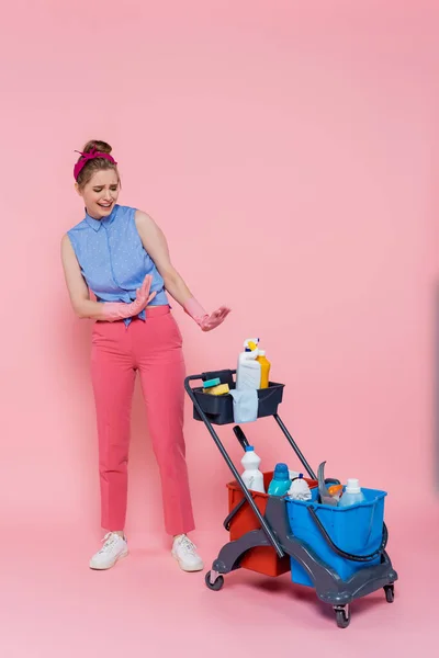 Comprimento total de chateado jovem mulher em luvas de borracha mostrando nenhum gesto perto de carrinho de limpeza em rosa — Fotografia de Stock