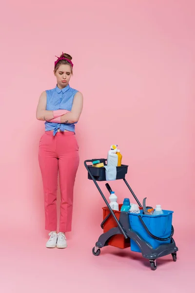 Pleine longueur de jeune femme offensée en gants de caoutchouc debout avec les bras croisés près du chariot d'entretien ménager sur rose — Photo de stock