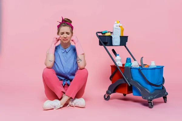 Pleine longueur de jeune femme bouleversée dans des gants en caoutchouc assis près du chariot d'entretien ménager avec des fournitures de nettoyage sur rose — Photo de stock