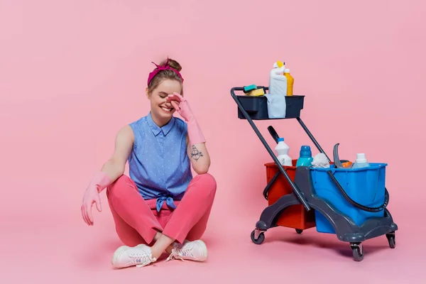 Comprimento total de sorrir jovem mulher em luvas de borracha sentado perto do carrinho de limpeza com suprimentos de limpeza em rosa — Fotografia de Stock