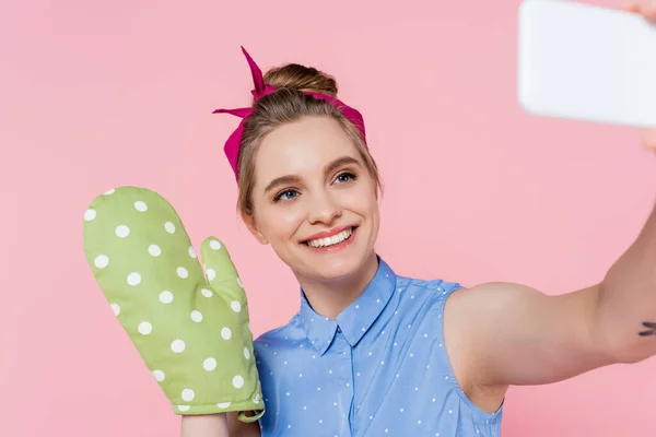 Щаслива молода жінка з зеленою рукавичкою для випічки бере селфі на смартфон ізольовано на рожевому — стокове фото