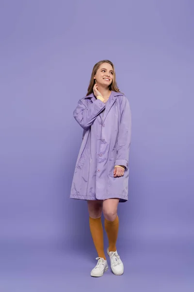 Повна довжина щасливої молодої жінки в фіолетовому дощовому пальто, що стоїть на фіолетовому — стокове фото