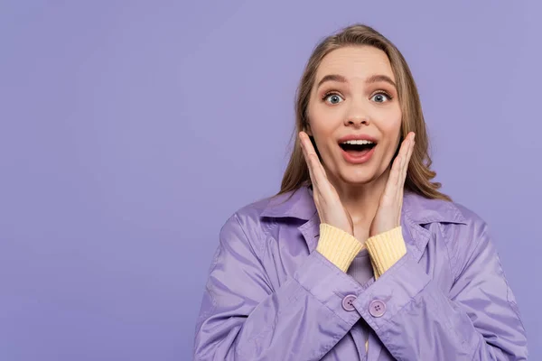 Mujer joven sorprendida en abrigo de lluvia violeta mirando a la cámara aislada en púrpura - foto de stock