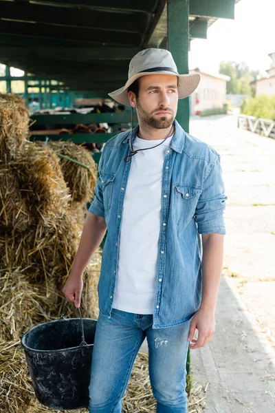 Joven agricultor en camisa de mezclilla y sombrero de ala sosteniendo cubo y mirando hacia otro lado en la granja - foto de stock