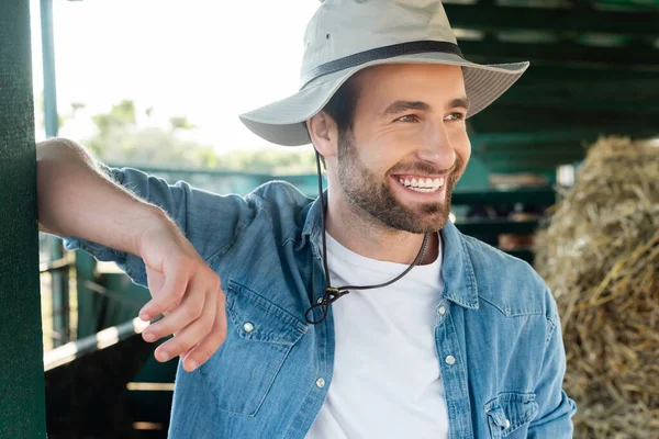 Agricultor alegre em chapéu de aba olhando para longe e sorrindo perto do celeiro — Fotografia de Stock