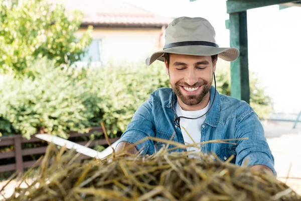 Jeune fermier barbu au chapeau de bord souriant près de la meule de foin à la ferme — Photo de stock