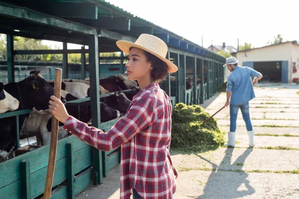 Joven afroamericana mujer mirando vaquero cerca granjero apilamiento heno en borrosa fondo - foto de stock