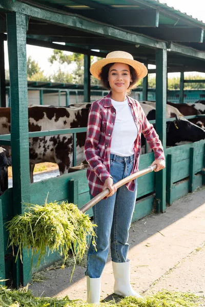 Happy african american woman looking at camera while stacking hay near cowshed — Stock Photo