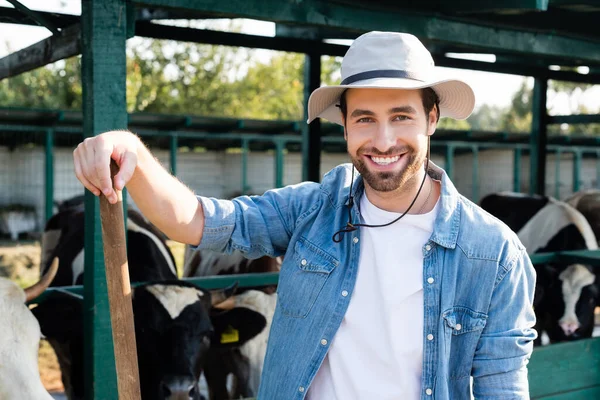 Cheerful farmer in brim hat smiling at camera near blurred cows — Stock Photo