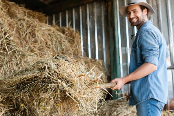 Granjero sonriente en sombrero de ala apilando heno sobre fondo borroso - foto de stock