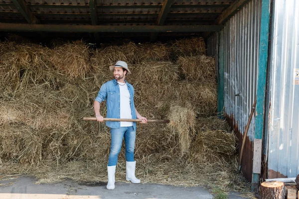 Вид на повну довжину щасливого фермера в джинсовому одязі і гумових черевиках біля стовбура сіна — стокове фото
