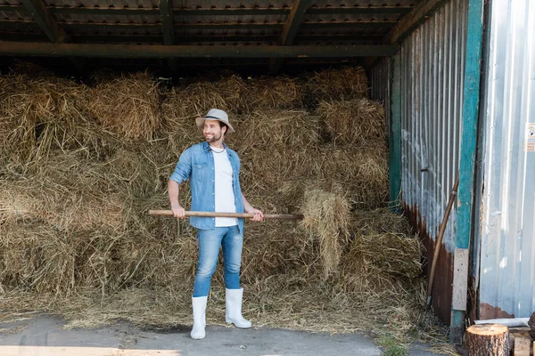 Vue pleine longueur de fermier heureux en vêtements en denim et bottes en caoutchouc près de la botte de foin — Photo de stock