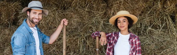 Cheerful interracial farmers in hats looking at camera near haystack, banner — Stock Photo