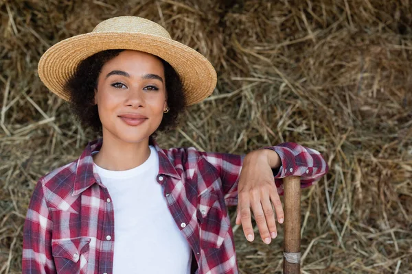 Positive african american woman in plaid shirt and straw hat smiling at camera near blurred haystack — Stock Photo