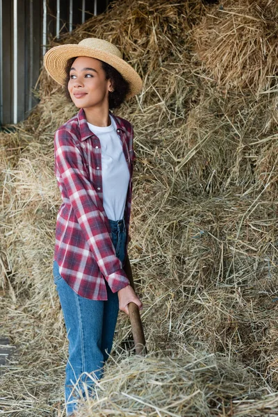 Mujer afroamericana feliz en camisa a cuadros y sombrero de paja mirando lejos cerca de pajar - foto de stock