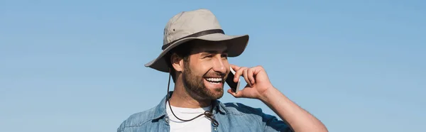 Granjero sonriente mirando hacia otro lado mientras habla en el teléfono móvil contra el cielo azul, bandera — Stock Photo