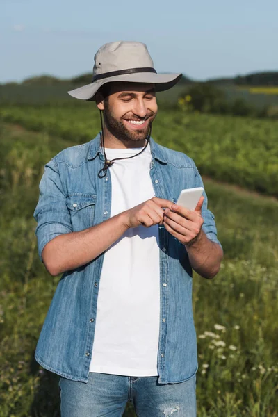 Agricultor feliz sombrero de ala y camisa de mezclilla de mensajería en el teléfono inteligente en el campo - foto de stock