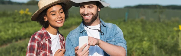 Jeune agriculteur montrant smartphone à un collègue afro-américain joyeux, bannière — Photo de stock