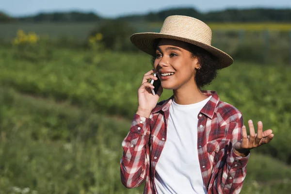 Sonriente mujer afroamericana en sombrero de paja hablando en el teléfono inteligente al aire libre - foto de stock