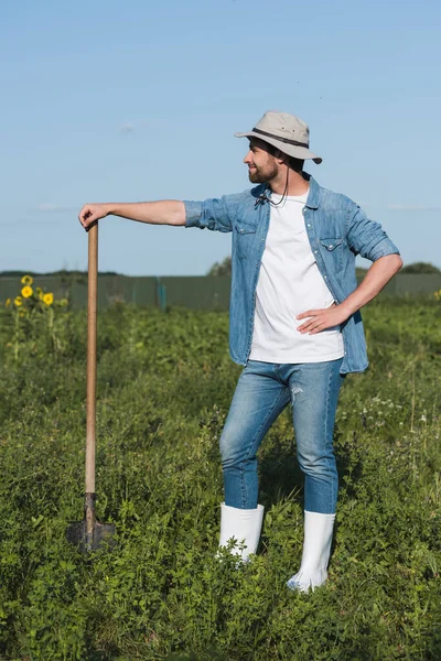 Piena vista di agricoltore felice con pala in piedi con mano sul fianco in campo — Foto stock